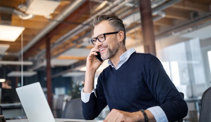 Uomo sorridente con il cellulare all’orecchio mentre guarda un computer portatile
